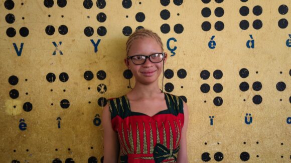 A young girl stands in front of a wall displaying a braille alphabet.