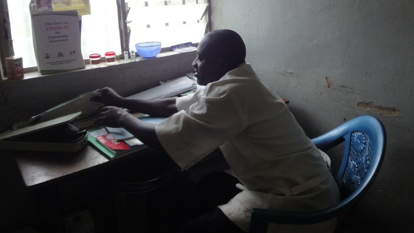 A man sits in a blue chair, next to a desk. The man was wearing a white shirt. The desk is piled high with books and folders.