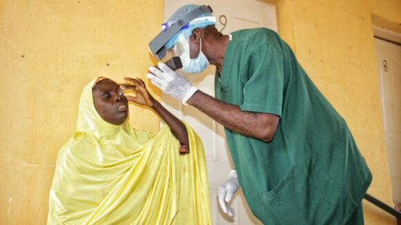 Zuwaira, a woman dressed in a yellow medical covering, having her eyes examined by an eye surgeon wearing PPE and green scrubs.