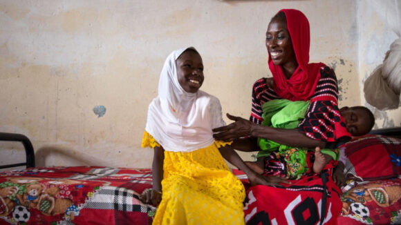 A girl and her mother sit on a bed, looking at each other and smiling.