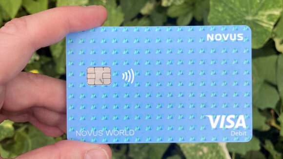 A hand holding a blue Novus bank card, featuring the text Visa and Novus World.