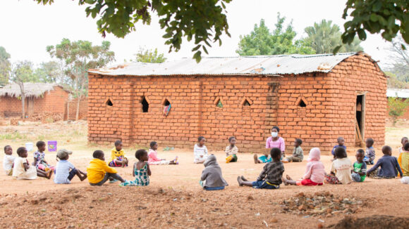 Young children sitting in a circle outside being taught vowels by their caregiver