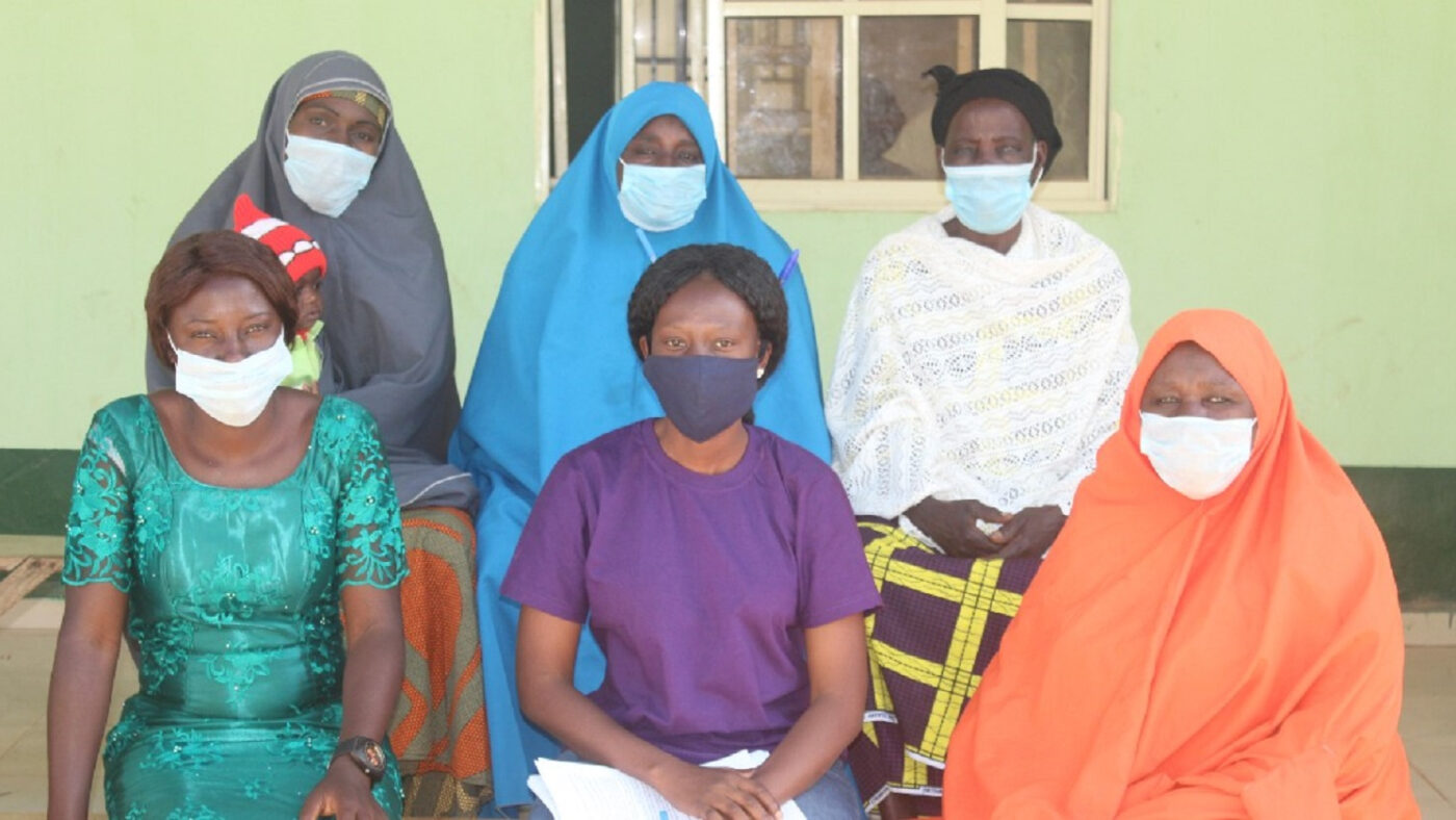 Six women sit down together and look at the camera. They are all wearing colourful headscarves, as well as facemasks.