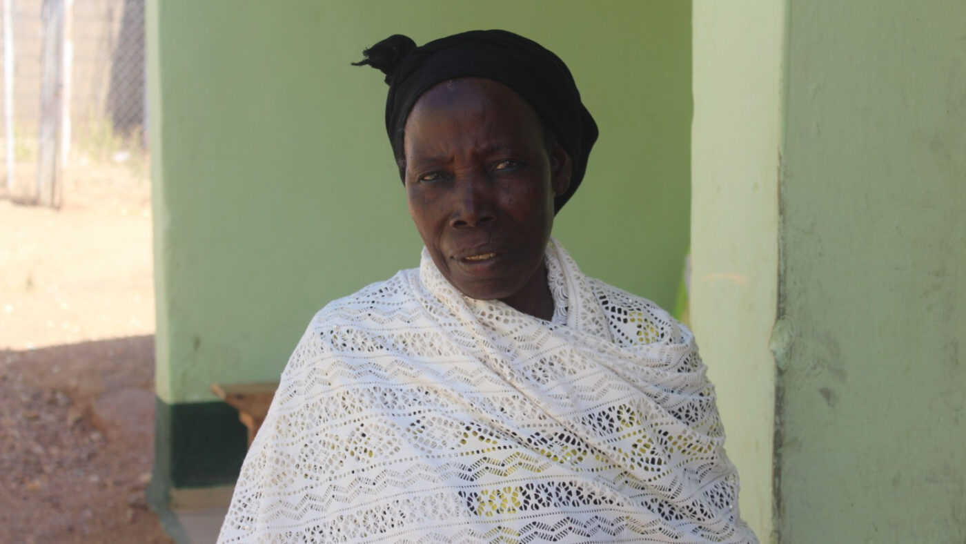 A woman sits down and looks at the camera. She is of African origin, wearing a white shawl, and has a black headscarf.