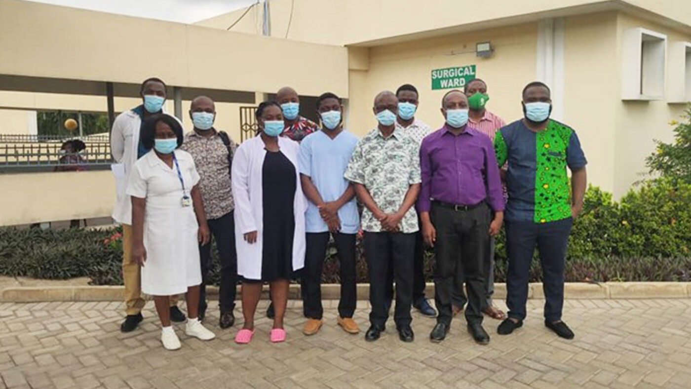 A group of health workers wearing surgical masks pose for a photograph in front of a hospital.