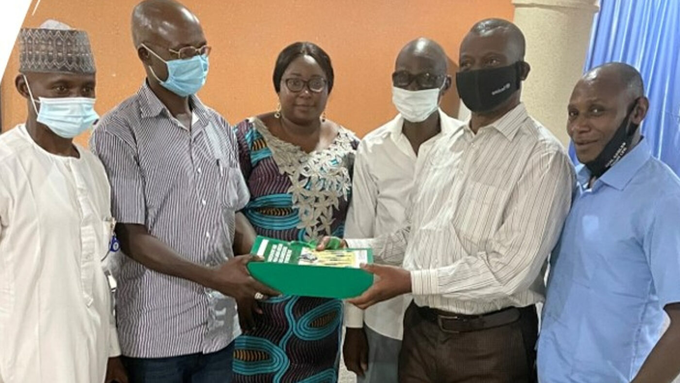 A group of men and women stand together, dressed smartly. One of the men is handing over a bundle of papers to another of the men.