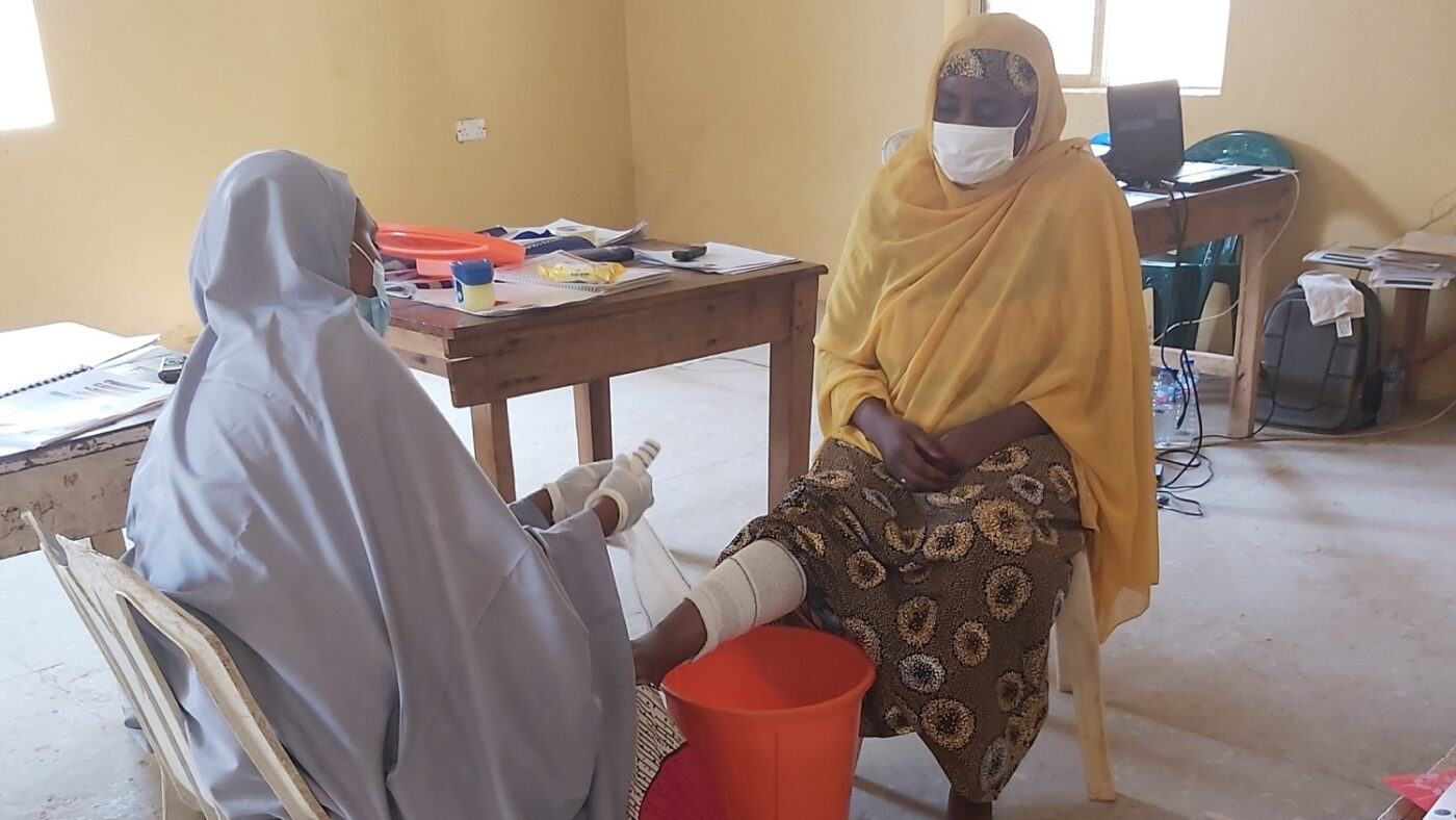 Two women sit opposite one another. One woman is holding the other woman's leg on her knee, and wrapping it in a white bandage.