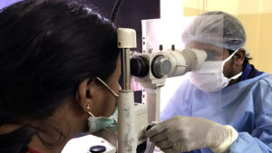 An eye health worker wearing a mask and surgical gown checks a woman's eyes using optical equipment.