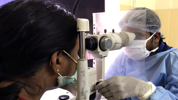 An eye health worker wearing a mask and surgical gown checks a woman's eyes using optical equipment.
