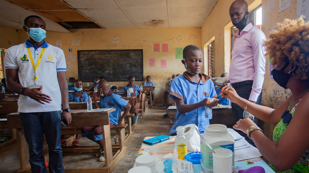 A school student in a classroom is handed a tablet by a health worker, as other health workers and his classmates look on.