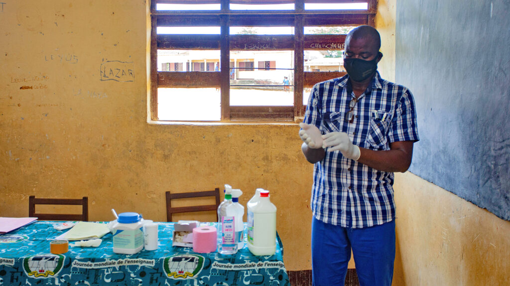 Teacher Biboum stands in his classroom in Cameroon, putting on gloves and preparing to give medication to students.