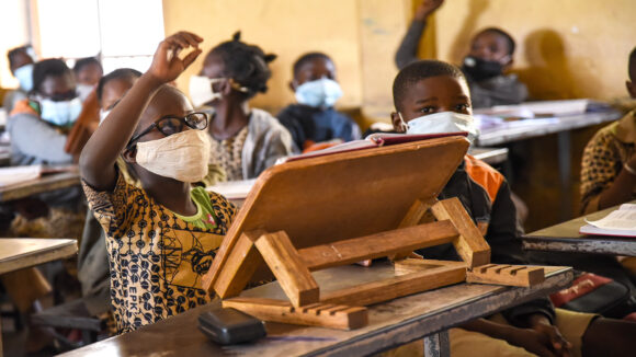 A school student raises her hand in class. She's wearing a mask and has an assistive lectern in front of her.