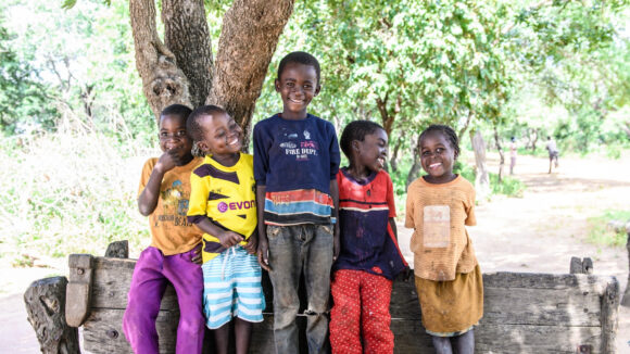 Five children smile and laugh, as they stand on a wooden bench with trees in the background.
