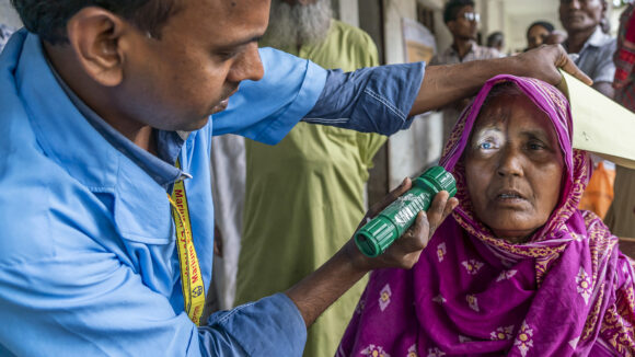 An eye health worker shines a torch into a woman's eyes to check for eye issues. She's wearing a colourful pink headscarf.