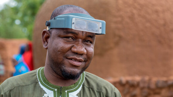 An eye health worker wearing magnifying glasses on his forehead. He's standing in a village with sandy-coloured huts behind him.