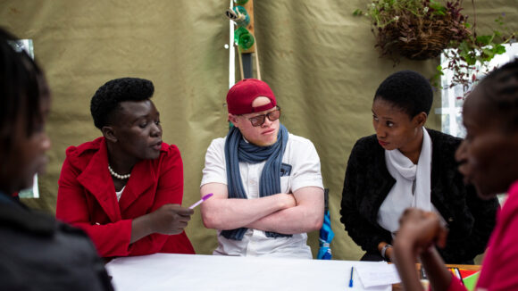 A group of young people sit around a table, talking together. The group includes both men and women, and most of the group members are wearing smart business clothes.
