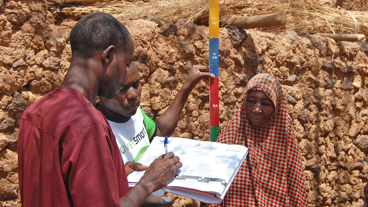 A community volunteer measures a woman using a dose pole to determine how much medication she needs. Another volunteer takes notes and records information.