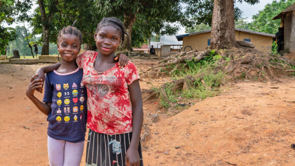 Two girls smile and link arms near their home, with dusty ground and trees in the background.