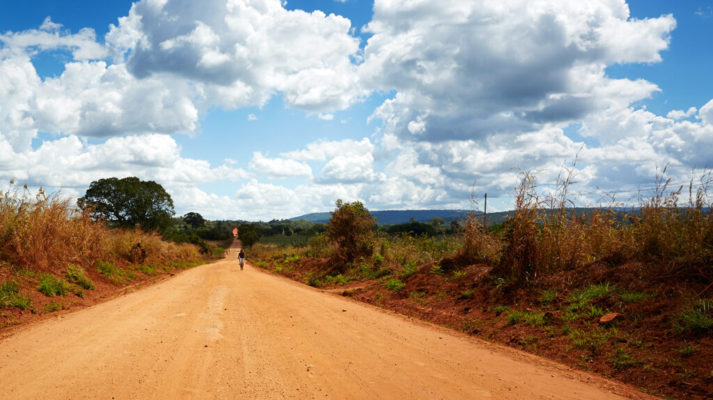 A dramatic Tanzanian landscape showing a red, dusty road stretching away into the distance, with greenery and mountains on the horizon.