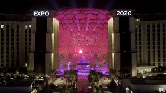 The Dubai Expo building, a huge glass dome lit up with pink lights and the words 'Expo 2020' illuminated above.
