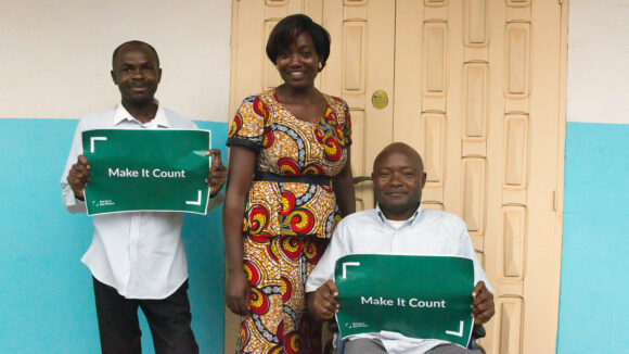 Two men and a woman in Cameroon. The men hold green posters saying 'Make it count'.