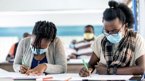 Two female students are sitting at a desk in a classroom, writing in notebooks. Shanice is sitting on the right.