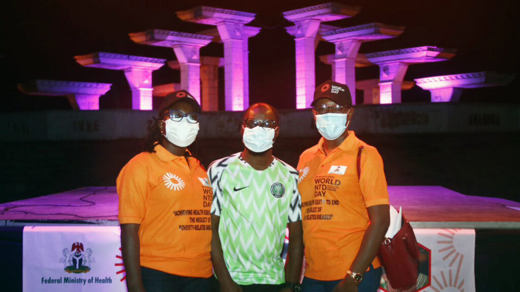 At night, three people stand together in front of Nigeria's Unity fountain, which is lit up in purple.