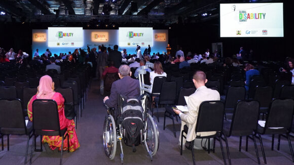 Audience members at the 2018 Global Disability Summit with their backs to the camera, looking at screens with the text 'Global Disability Summit'.