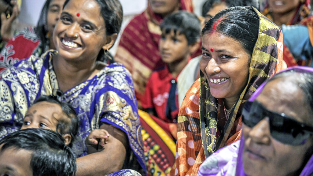 A group of women wearing colourful headscarves smile and laugh during a puppet show in their community.
