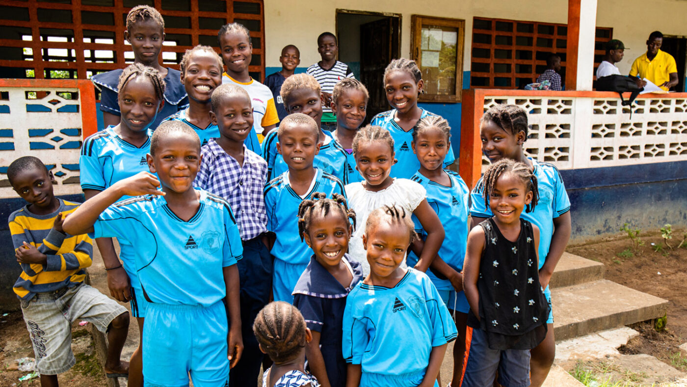 A large group of smiling children gather for a group photo outside their school.