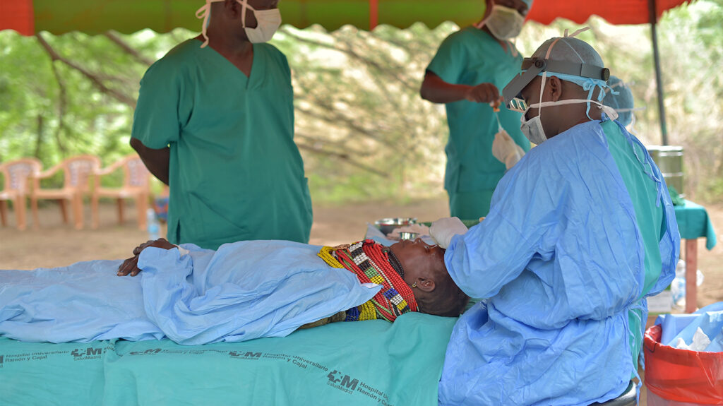 Dressed in surgical scrubs, Samson performs trachoma surgery on a woman outside under a canopy. Two male eye health workers, who are also dressed in surgical scrubs, are on hand to assist.