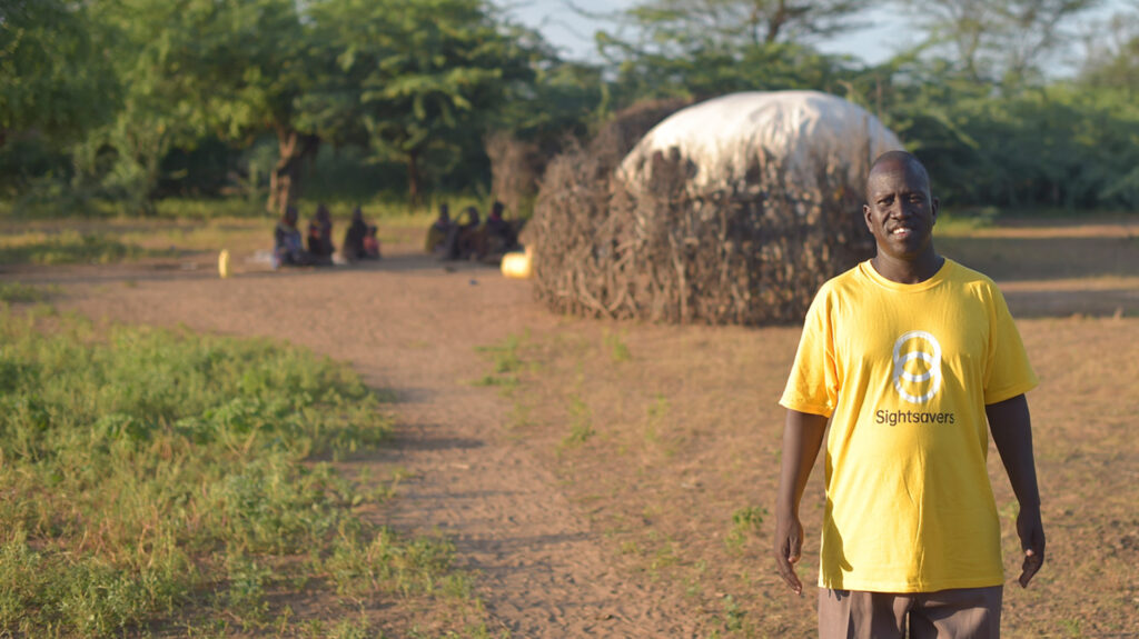 Samson is standing in a desert environment, wearing a yellow Sightsavers t-shirt. In the background, a circle of people are sitting in a circle outside a tent.