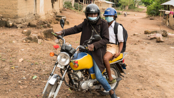 A man sits on a stationary motorbike with Saio on the backseat. They are both wearing helmets and surgical masks.