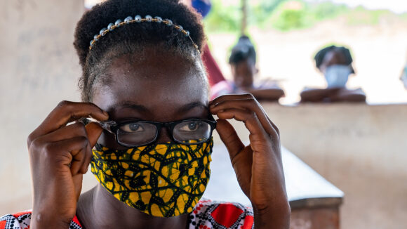 A girl wearing a cloth face mask adjusts her glasses to read a vision chart.