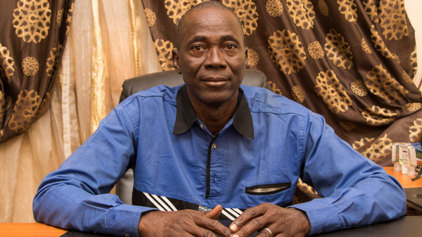 Dr Yocuba Sangare sits in an office chair, with a neutral expression. Behind him, there are ornate curtains and drapes.