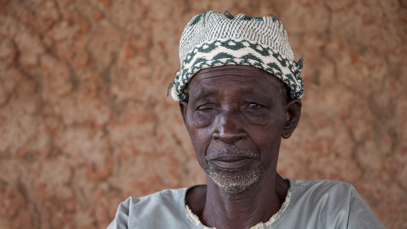 Mamourou sits with a mud wall behind him. His left eye is partially closed.