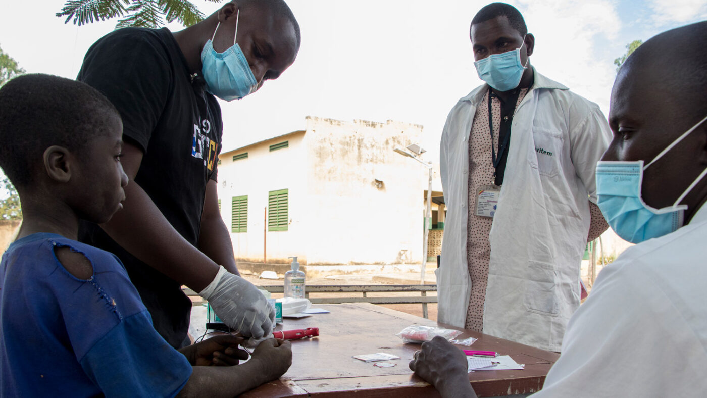 Lassina gives a blood spot sample at school while three medical professionals wearing surgical masks look on.