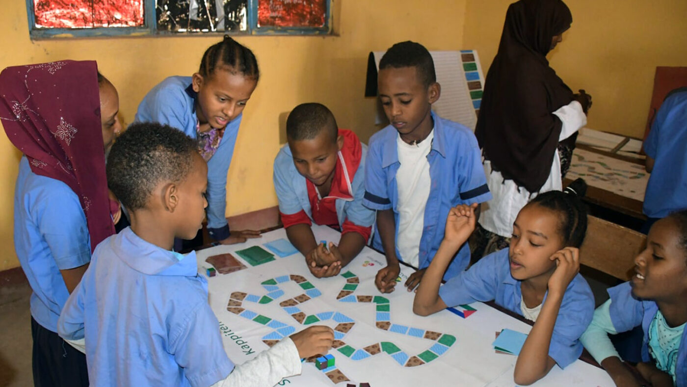 A group of seven schoolchildren gather around a table where they are playing a board game.