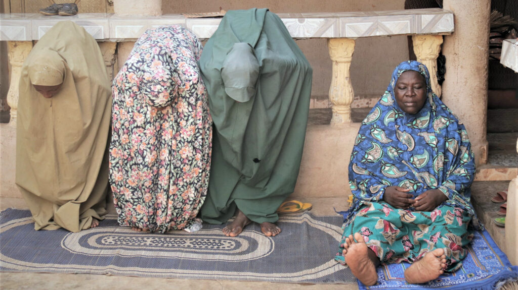 Salifat sits on a mat while praying. Next to her, three women are bending over to pray.