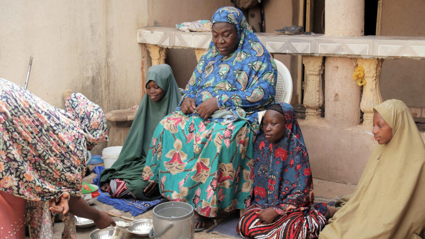 Salifat sits in a chair outside her home while her granddaughters sit around her on the ground. A woman is bending over to serve white rice into silver bowls.