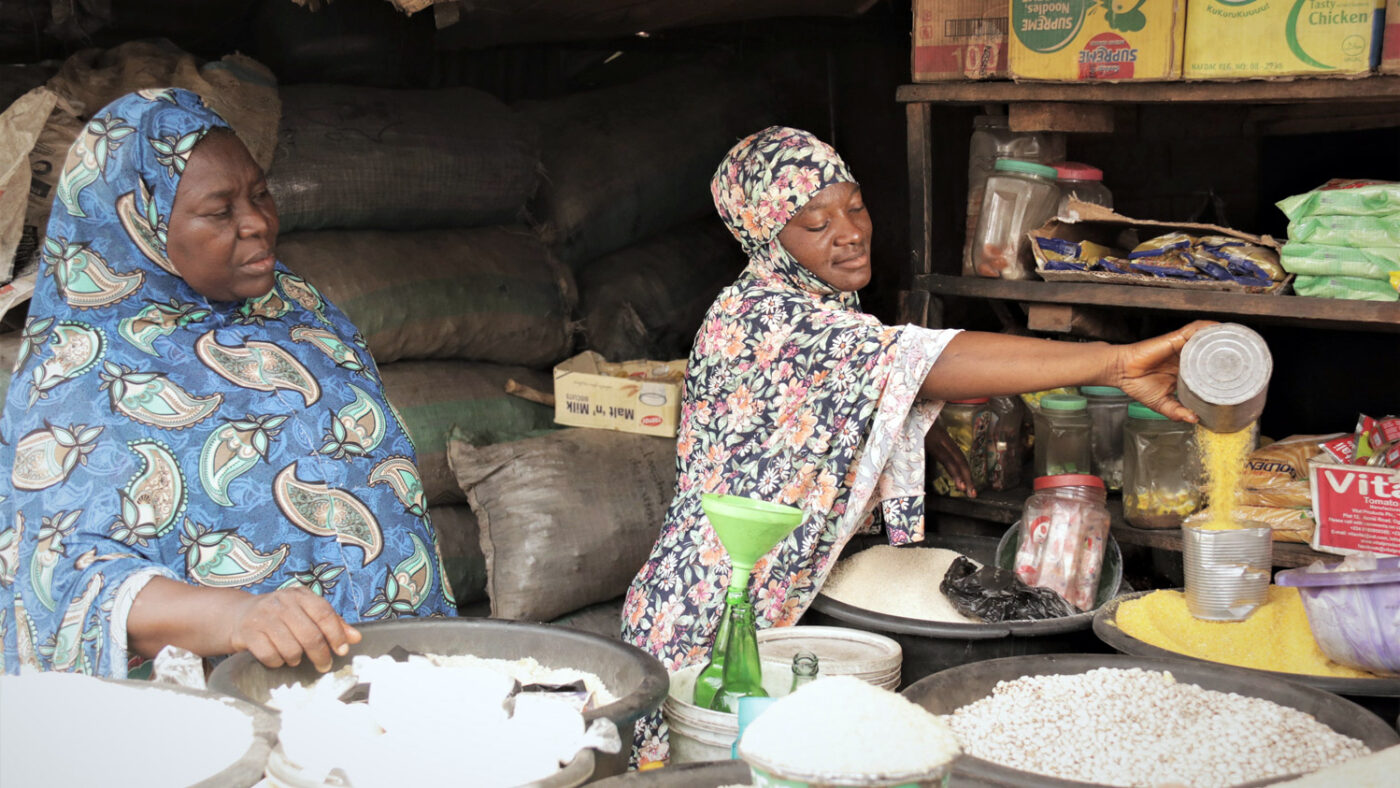 Salifat watches her daughter Mainna pour lentils into a container. They are standing in front of bowls of rice and pulses.