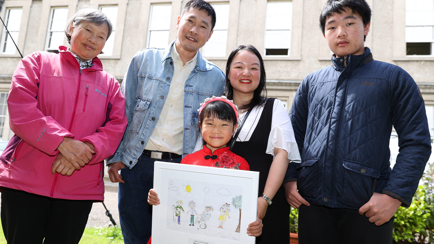 Quinglan smiles while holding her framed drawing, which depicts four people, two of whom have a disability. Quinglan is joined by four members of her family, who are standing behind her smiling.