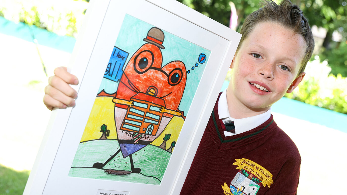 Tom stands smiling while holding his framed drawing. The drawing depicts a character shaped like a heart holding a building, which looks like a hospital. Some text on the drawing reads 'I love all'.