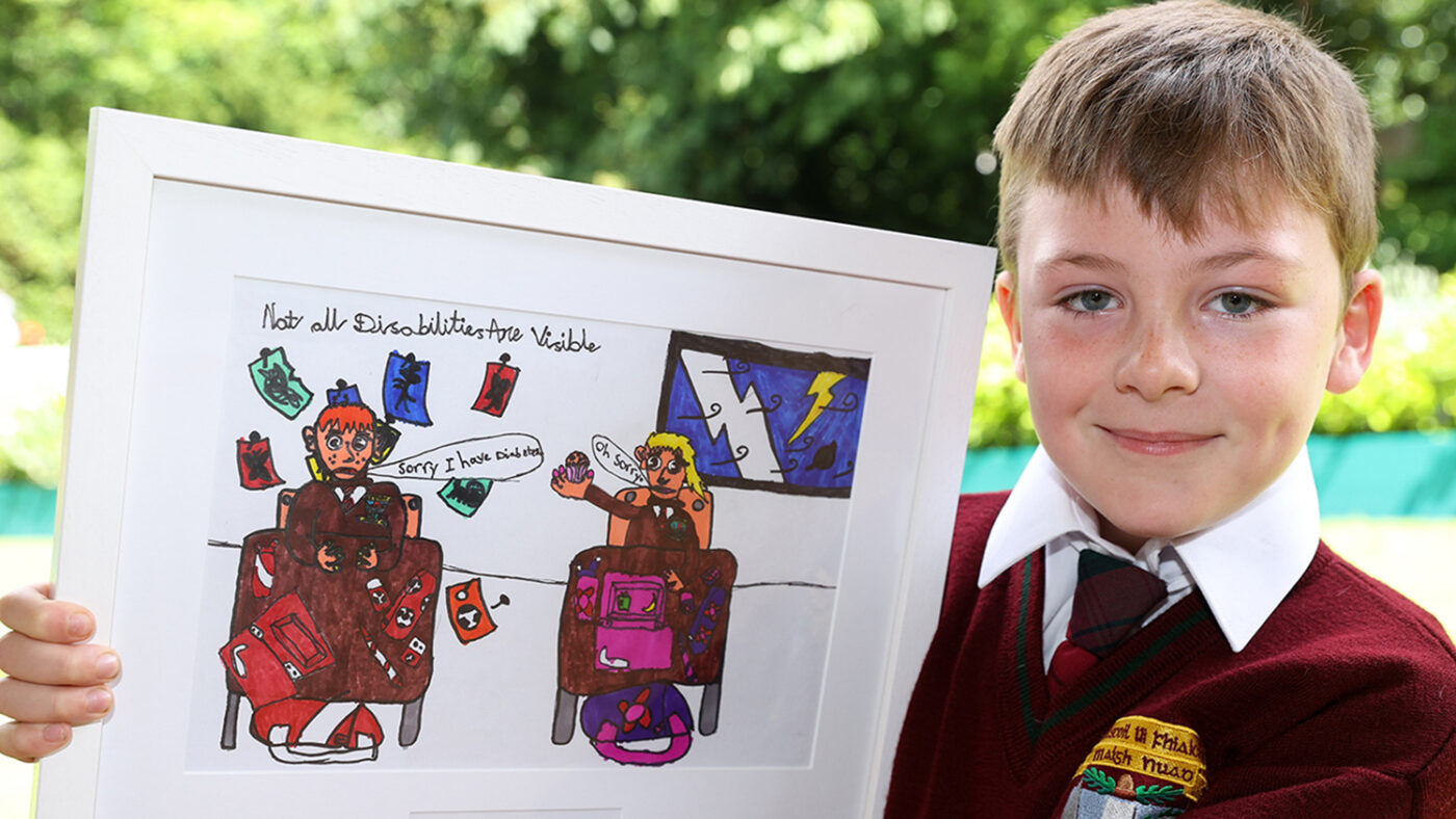 Dylan holds his framed drawing, which shows two pupils sitting at desk having a conversation. One of them shares that they have a disability. The text on the drawing reads 'Not all disabilities are visible'.