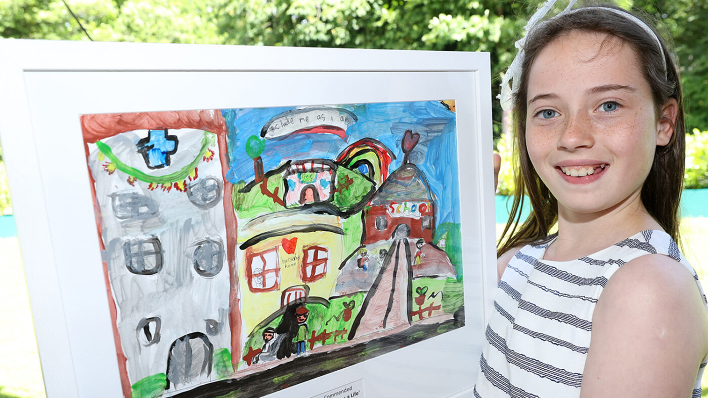 Kate smiles and holds her painting, which shows a community including a school and a nursing home. The text on the painting reads 'Include me as I am'.