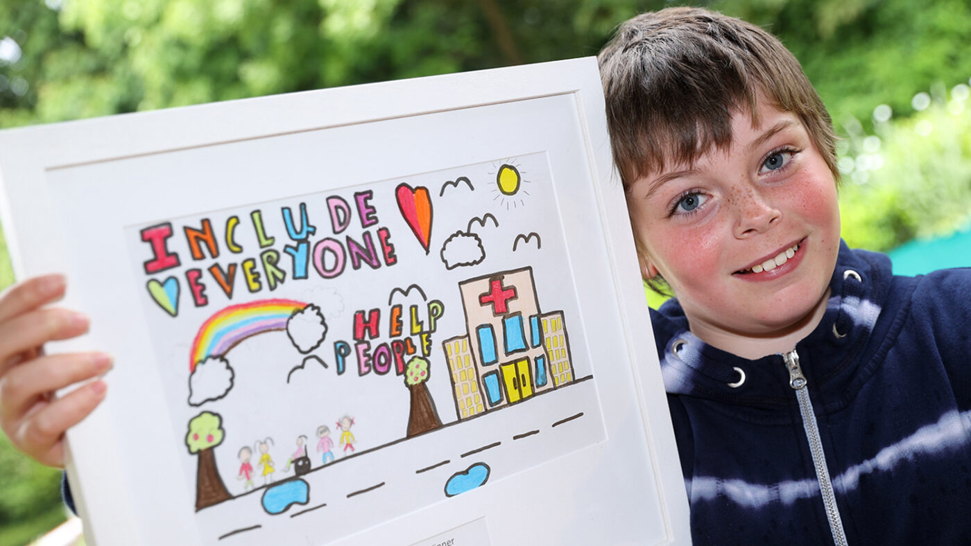 Lyla smiles while holding their colorful, framed drawing. The drawing shows a hospital and group of people, one of whom is using a wheelchair. The text reads Include everyone. Help people'.