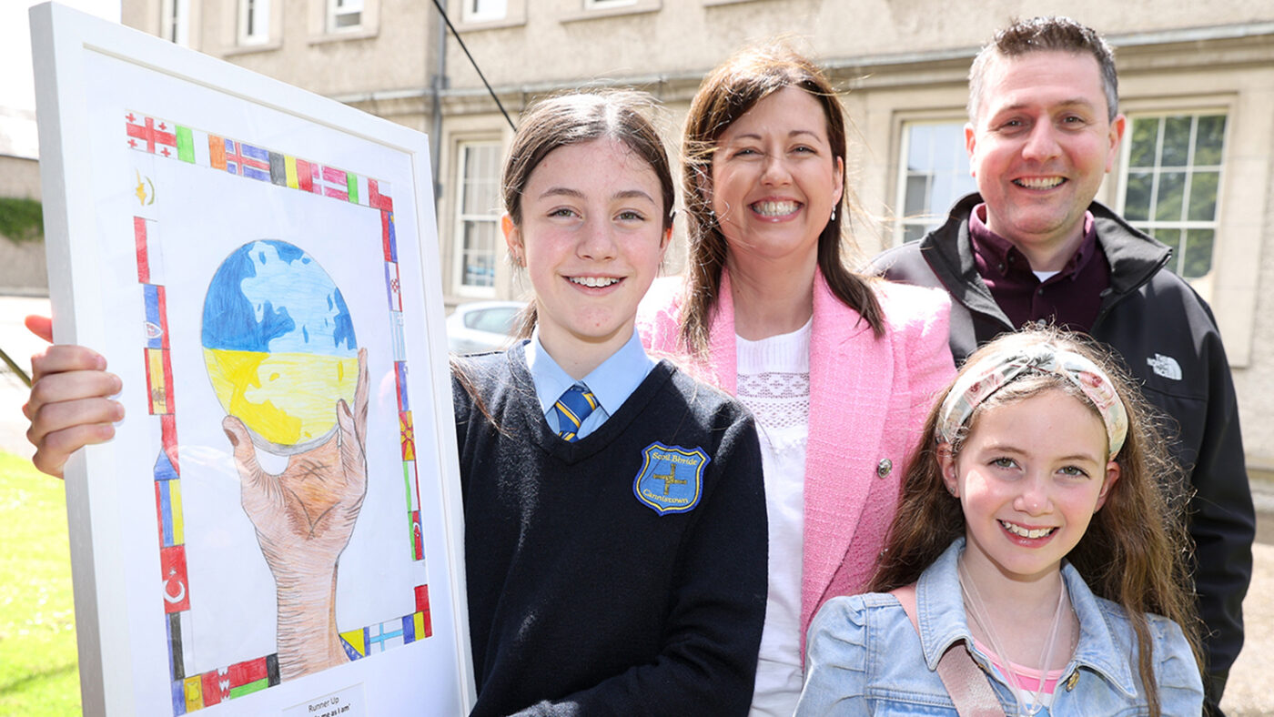 Brooke, her parents and younger sister smile while she holds her framed drawing. The drawing depicts a hand holding a globe, surrounded by a border of world flags.