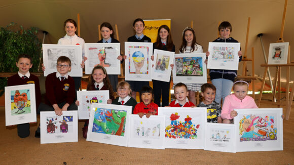 A large group of primary school children smile and pose for a photo as they hold their artwork.