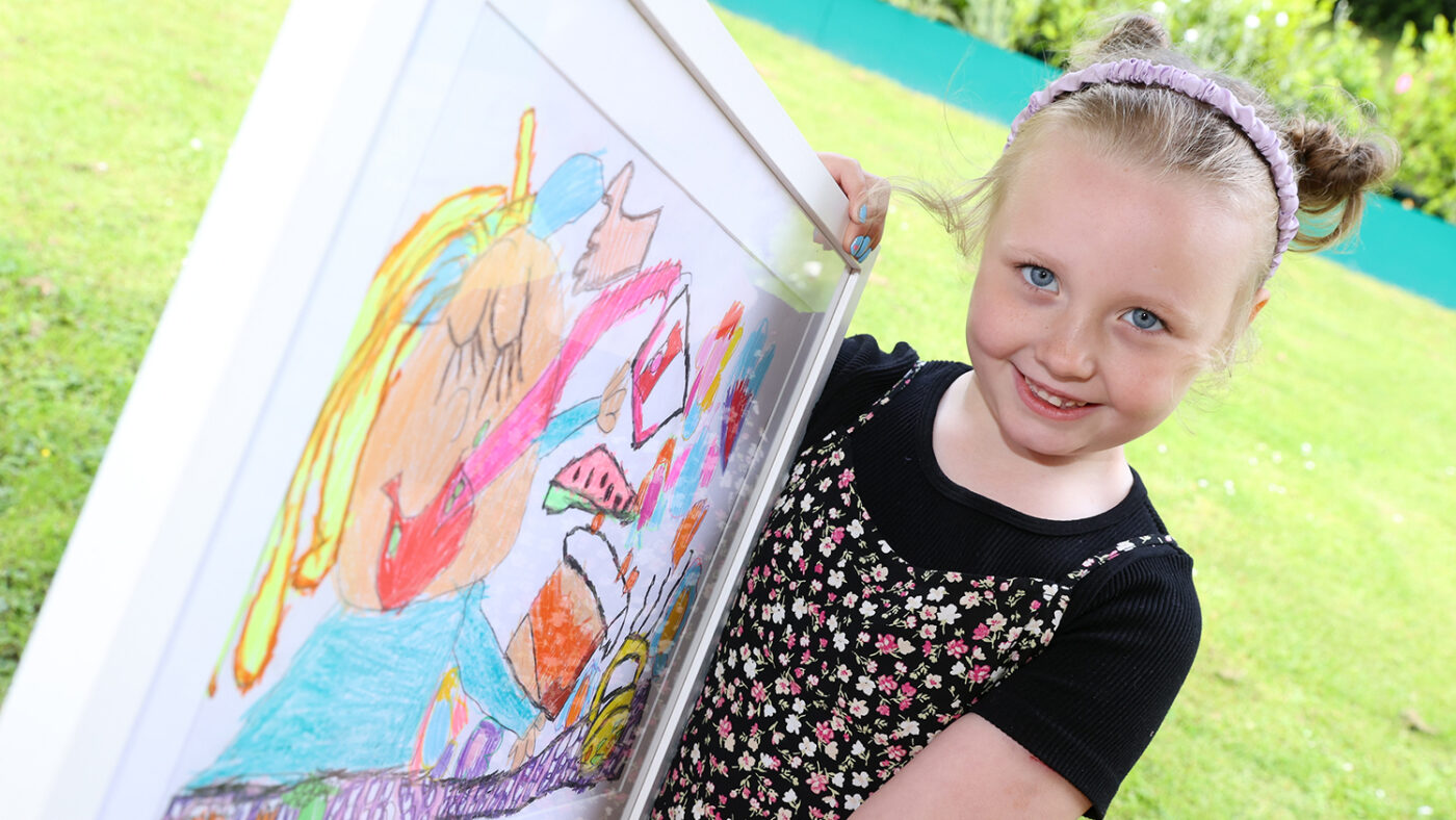 Ella smiles while holding her framed drawining, which shows a girl eating and enjoying various foods.
