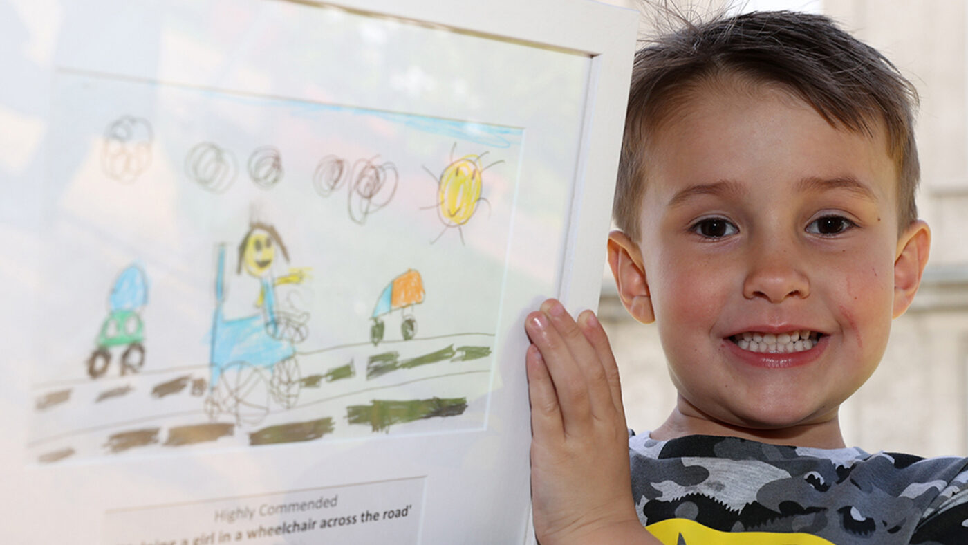 Jack smiles while holding his framed drawing of a girl in a wheelchair crossing the road.